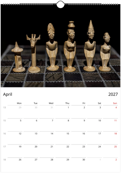 A wall calendar page showing the month of April 2027 below a large photograph of six carved wooden figurines on a dark grid surface. The calendar has a white lower section with black text and a small red day highlight for the 4th.