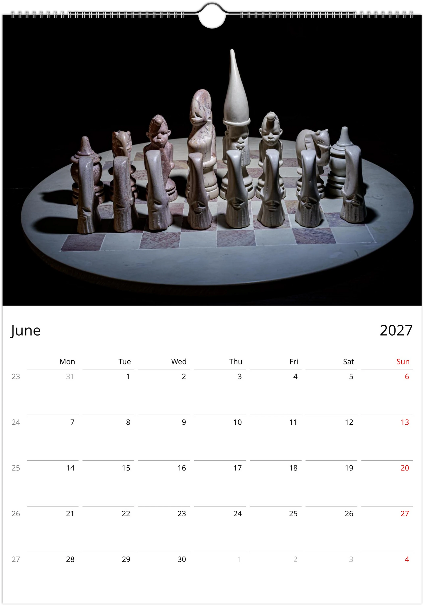 A spiral-bound wall calendar page showing a photograph of a sculptural chess set on a circular table against a black background, with a June 2027 calendar grid below.