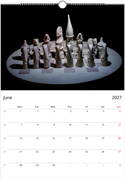 A spiral-bound wall calendar page showing a photograph of a sculptural chess set on a circular table against a black background, with a June 2027 calendar grid below.