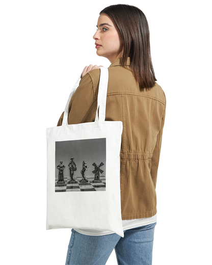 A woman wearing a brown jacket and blue jeans is shown from the back, carrying a white canvas tote bag over her shoulder. The tote bag features a black and white graphic print of chess pieces on a chessboard.