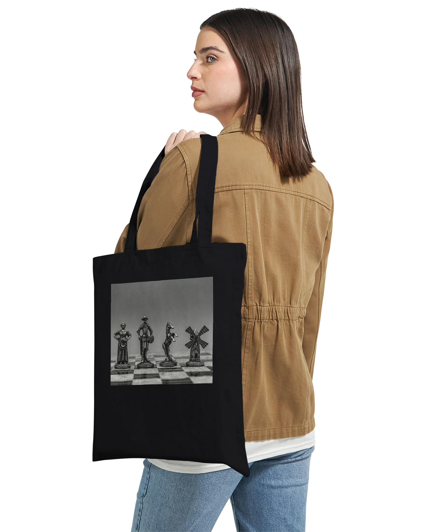 A woman wearing a tan jacket and blue jeans carries a black canvas tote bag over her shoulder. The bag features a square black-and-white graphic print of chess pieces on a chessboard.