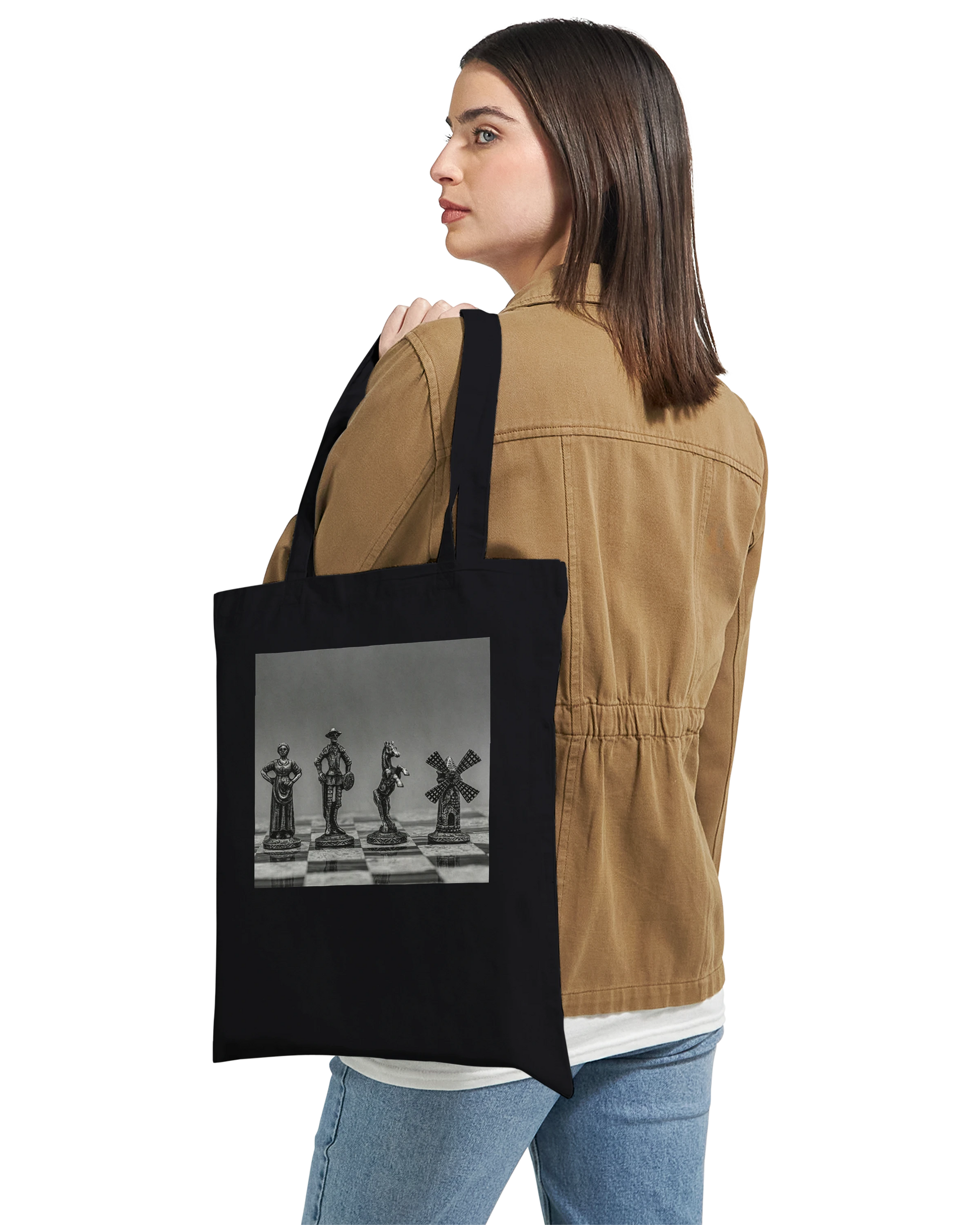 A woman wearing a tan jacket and blue jeans carries a black canvas tote bag over her shoulder. The bag features a square black-and-white graphic print of chess pieces on a chessboard.