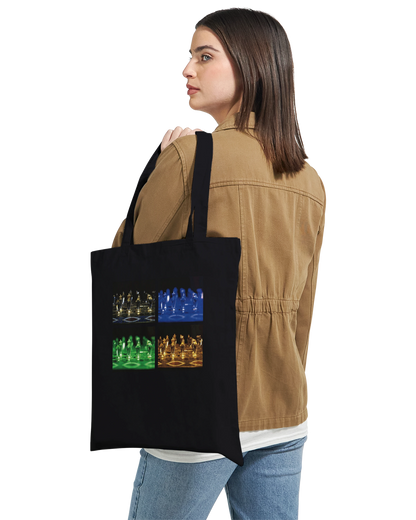 A woman wearing a tan jacket and blue jeans carries a black canvas tote bag over her shoulder. The bag features a graphic print of four chess-themed panels in blue, green, and orange.