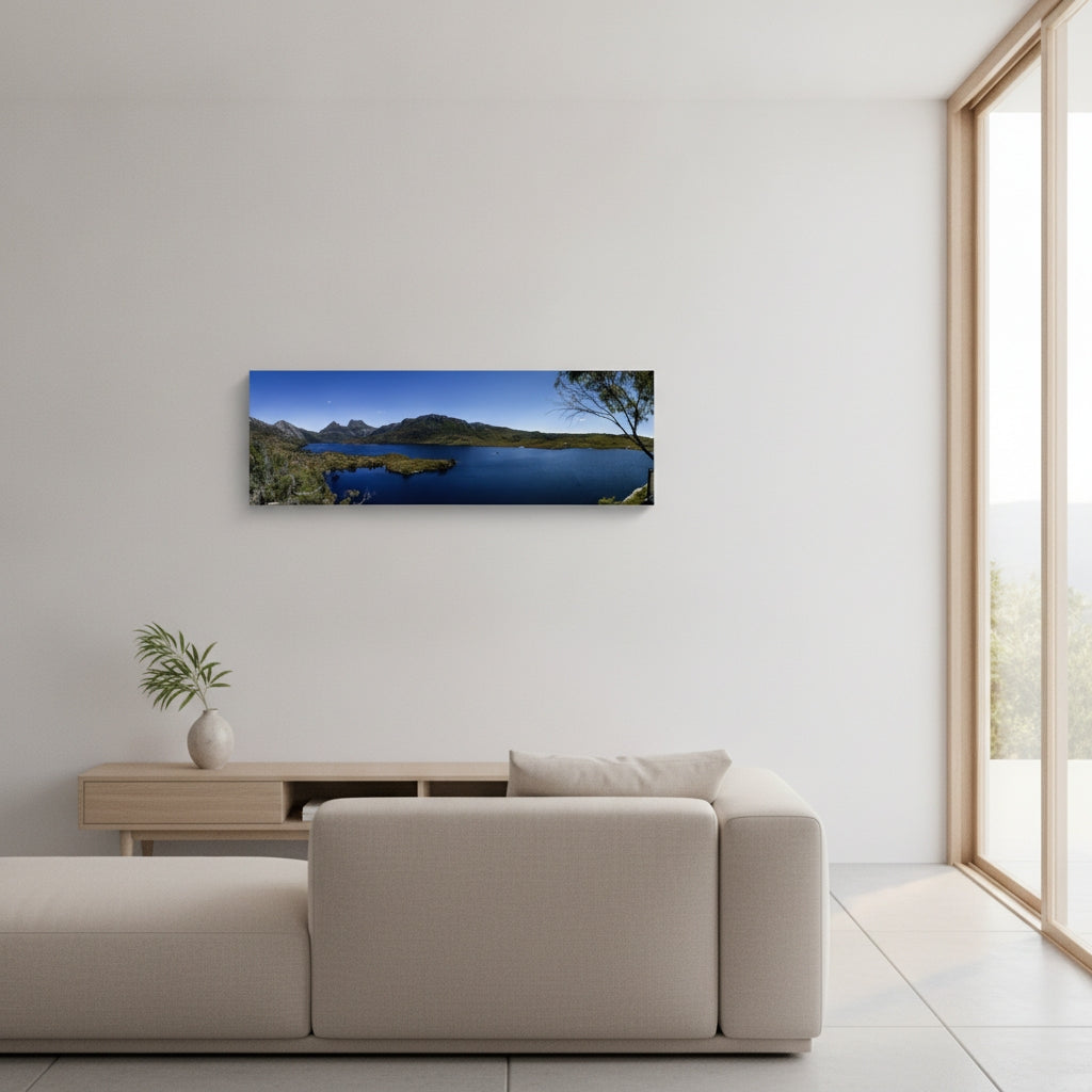 A panoramic landscape wall art print of a blue lake and Cradle Mountain, displayed on a white wall in a modern living room setting above a beige sofa.